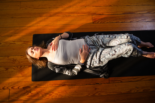 Picture of a female laying down on a mat on a wood 
         floor, her left hand on her chest and right hand on her stomach